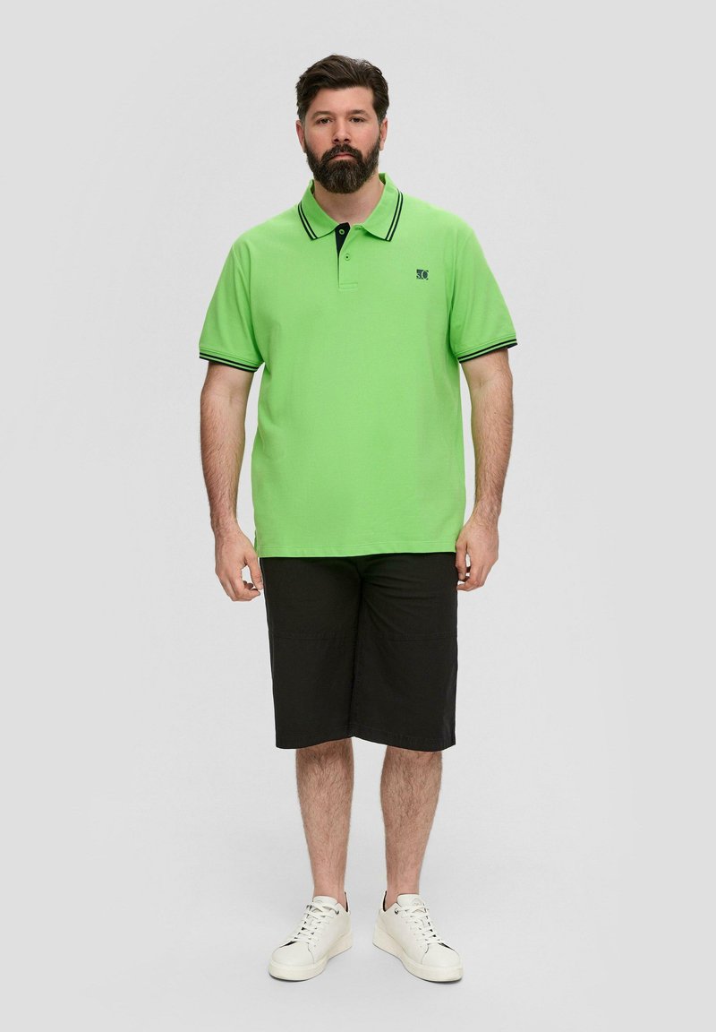 Zielona koszulka polo z czarnymi akcentami, zestawiona z czarnymi szortami i białymi sneakersami. Standardowy krój z kołnierzykiem i zapięciem na dwa guziki.