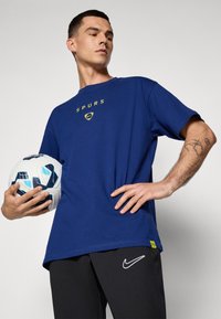 Blå bomulds T-shirt med "SPURS" logo i gul, båret med sorte Nike bukser. Holder en hvid fodbold med blå accenter.