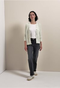 Hellgrüne Strickjacke mit vertikalen Rippen, weißes T-Shirt, dunkelgraue Jeans und weiße Sneakers mit einer beigen Sohle, stehend vor einem neutralen Hintergrund.