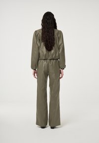 Vrouw met lang, krullend haar, gekleed in een olijfgroene, glanzende jas en wijde broek, staande met haar rug naar de camera op een effen achtergrond.