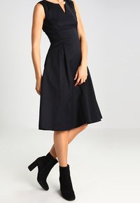Robe midi noire sans manches avec un décolleté en V et une jupe plissée. Portée avec des bottines noires en daim au niveau de la cheville avec un talon épais. Texture lisse.