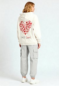 Felpa crema con motivo a cuori rossi sul retro, con la parola "SWEET" e "heart" sottostante. Indossata con pantaloni cargo grigi e scarpe da ginnastica bianche.