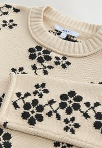 Pull en maille beige avec un motif floral noir, présentant un col rond côtelé et un tissu texturé. Étiquette visible au niveau du col.