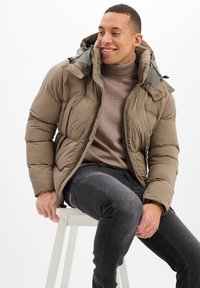 Chaqueta acolchada marrón con cierre de cremallera, capucha ajustable y bolsillos laterales, combinada con un cuello de tortuga beige y jeans oscuros.