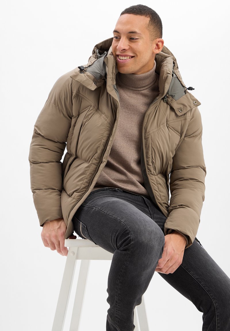 Braune Pufferjacke mit Reißverschluss, verstellbarem Kapuzen, und Seitentaschen, kombiniert mit einem beigen Rollkragenpullover und dunklen Jeans.