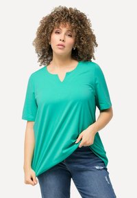 Maglietta turchese a maniche corte con scollatura a V, realizzata in tessuto morbido, caratterizzata da una vestibilità rilassata e un leggero drappeggio. Indossata con jeans blu strappati.
