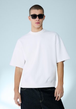 Nuori mies, jolla on mustat aurinkolasit, ylisuuri valkoinen lyhythihainen paita ja tummat housut vaaleansinisen taustan edessä.