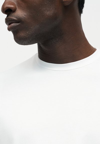 Κοντινό πλάνο στο κάτω μέρος του προσώπου και τον λαιμό ενός ατόμου με σκούρο δέρμα, που φοράει απλό άσπρο μπλουζάκι με στρογγυλή λαιμόκοψη με φόντο λευκό.