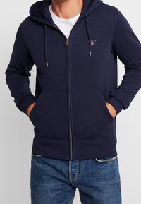 Sweat à capuche zippé bleu marine en mélange de coton lisse, doté d'une capuche avec cordon de serrage, de poches avant, de poignets côtelés et d'un petit logo sur la poitrine.