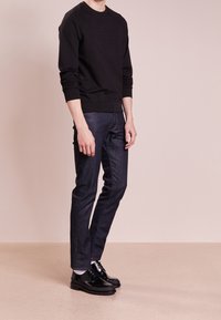 Černá mikina s vyraženým logem, v kombinaci s tmavě modrými slim-fit džíny a černými šněrovacími botami. Jednoduché pozadí, bez v�ýrazných textur.