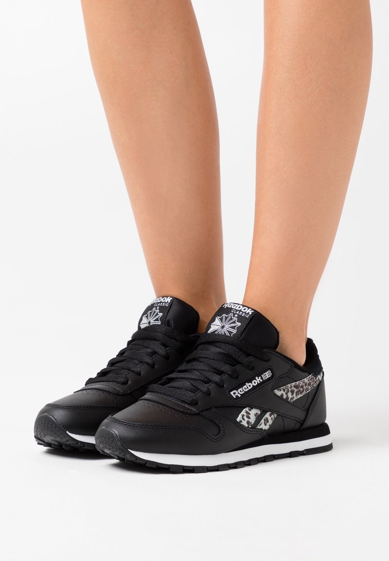 Jambes portant des baskets Reebok Classic noires avec semelles blanches et détails à imprimé léopard sur les côtés, sur fond blanc.