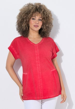 Kvinde med krøllet hår iført en rød kortærmet V-hals top og hvide bukser, der står mod en ensartet lys baggrund.