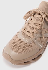 Scarpa sportiva beige con parte superiore in mesh, accenti testurizzati e lacci rotondi. Presenta un colletto imbottito e un design stratificato con una suola affusolata.