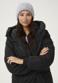 Black puffer coat with a large hood, featuring a zip front and ribbed cuffs. Gray knitted beanie, long dark hair, and crossed arms.