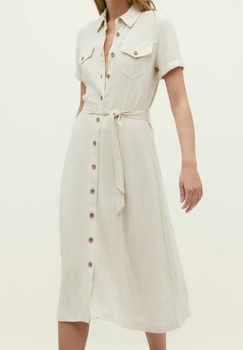 Robe midi beige à boutons, manches courtes, poches poitrine et ceinture nouée à la taille, portée par une femme debout sur un fond uni.