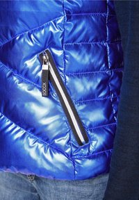 Blauer Puffermantel mit glänzender, gesteppten Textur, ausgestattet mit einer schwarzen, mit Reißverschluss versehenen Tasche mit einem weißen Streifenakzent auf der Vorderseite.