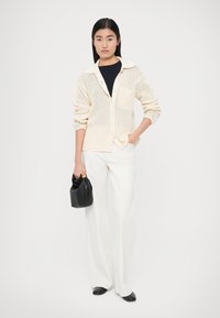 Πλεκτή ζακέτα σε εκρού χρώμα με γιακά, κουμπιά στο μπροστινό μέρος και τσέπη. Συνδυασμένη με λευκό παντελόνι φαρδιάς γραμμής και μαύρη τσάντα.