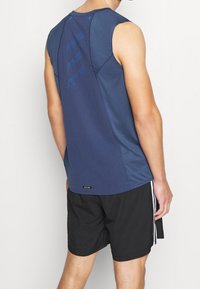 Námořnicky modrý sportovní top bez rukávů s texturovanou síťovanou látkou, s geometrickým vzorem na zádech a kontrastními modrými akcenty. Černé šortky.