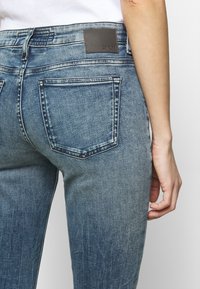 Primer plano de una persona que lleva jeans azul desteñidos, destacando los bolsillos traseros y la etiqueta de marca de cuero en la cintura.