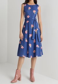 Blå blommig klänning med en figursydd bodice, ärmlös design och knytning i midjan. Har rosa blommönster och når under knäet. Rosa ankelskor.