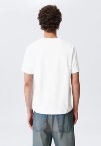 Weißes Baumwoll-T-Shirt mit entspannter Passform, kurzen Ärmeln und Rundhalsausschnitt. Kombiniert mit hellblauen Jeans aus Denim mit Gesäßtaschen.