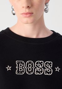 Felpa nera con logo "BOSS" ricamato in bianco testurizzato e dettagli a forma di stella. Collo a costine. Orecchini a bottone bianchi sono visibili.