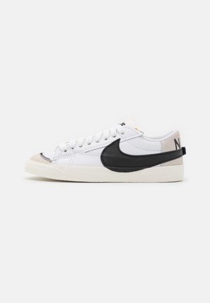 Biały niski sneaker z czarnym logo Nike swoosh, beżowym noskiem, białymi sznurówkami i kremową podeszwą na jednolitym tle.