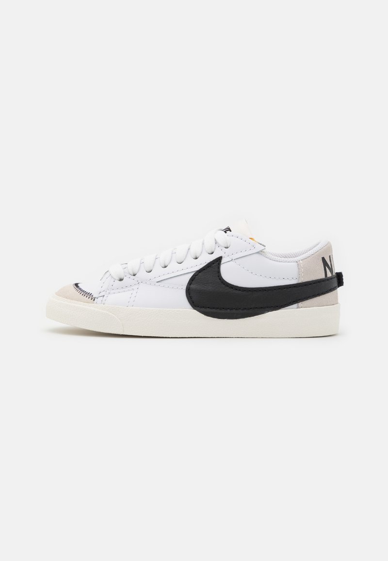 Basket basse blanche avec le logo Nike swoosh noir, embout beige, lacets blancs et semelle blanc cassé sur un fond uni.