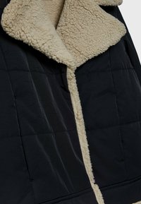 Veste matelassée noire avec doublure en sherpa beige duveteuse visible le long du col et des bords avant.