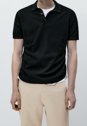 Czarna koszulka polo z fakturowanym splotem, krótki rękaw i klasyczny kołnierzyk, zestawiona z jasnobeżowymi spodniami.