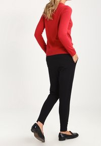Pull rouge à manches longues associé à un pantalon noir fuselé. La tenue présente une texture lisse et des chaussures plates noires. Design minimaliste.