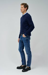 Pull marin sur chemise bleu clair, jean bleu et mocassins noirs. Le pull a des poignets côtelés. Jean droit avec un léger effet usé.