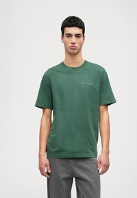 Zaļš kokvilnas T-krekls ar īsām piedurknēm un apaļu kakla izgriezumu, ar smalku tekstu uz krūtīm, apvienots ar pelēkiem džinsa biksēm.