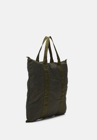 Sac en nylon vert avec un design matelassé, doté de deux poignées en tissu et d'une fermeture éclair sur le côté. Texture lisse et accents minimalistes.