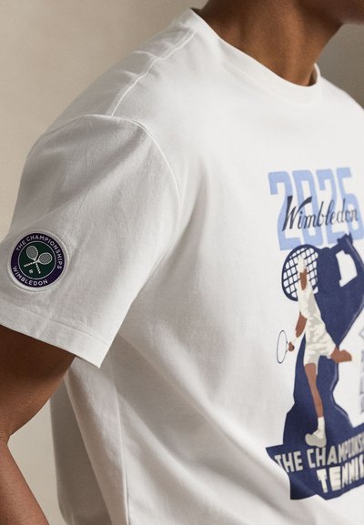 Λευκό μπλουζάκι με το λογότυπο του Wimbledon 2025 και γραφικό ενός τενίστα που χτυπάει τη μπάλα, με ραμμένο έμβλημα των Πρωταθλημάτων Wimbledon στο μανίκι.
