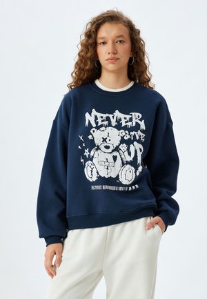 Marineblauw sweatshirt met een groot wit graphic van een teddybeer en de tekst "GEEN OPGEVEN." Voorzien van ribbels aan de mouwen en onderkant, met een relaxte pasvorm.