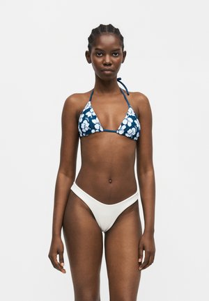 Fonott hajú nő, kék virágmintás háromszög bikini felsővel és sima fehér bikini alsóval, áll egy világos, egyszínű háttér előtt.