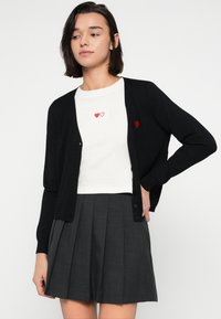 Μαύρο ζακέτα από απαλό ύφασμα, με κούμπωμα με κουμπιά και κέντημα κόκκινης καρδιάς στο αριστερό στήθος. Συνδυασμένο με λευκό τοπ και γκρι πλισέ φούστα.