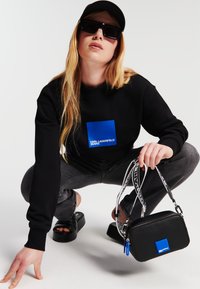 Sudadera negra con un logotipo cuadrado azul, jeans grises, sandalias de plataforma negras y un bolso pequeño negro con un acento azul y logotipo.