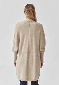 Cardigan en tricot beige, texture douce, coupe oversize, poignets côtelés, détail de couture au dos et ourlet arrondi. Design décontracté adapté pour les superpositions.