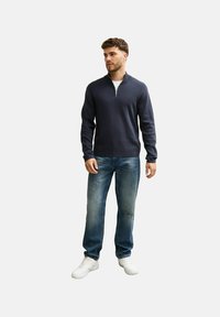 Himmelblauer Half-Zip-Pullover aus weichem Material, kombiniert mit ausgebleichten blauen Jeans und weißen Sneakern, auf einem neutralen Hintergrund präsentiert.