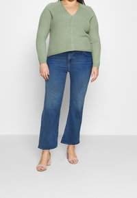 Gilet côtelé vert menthe avec décolleté en V et fermeture à boutons, associé à un jean en denim bleu évasé et des sandales à talons blocs couleur rose blush.