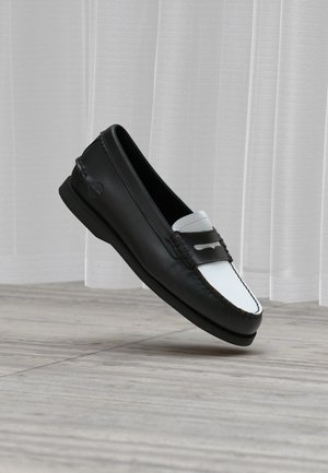Zwart-witte leren loafer met moc-toenaad en decoratieve band, zwevend boven een lichte houten vloer en doorschijnende gordijnen afgebeeld.