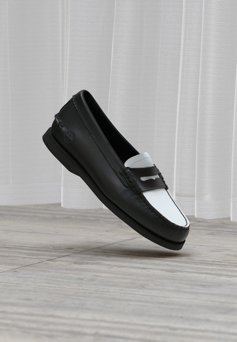 Mocassin en cuir noir et blanc avec couture moc toe et bride décorative, présenté flottant au-dessus d’un sol en bois clair et de rideaux légers.