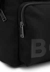 Sac à dos noir en nylon avec des compartiments zippés et un logo en relief bien visible. Surface texturée et accents en matériel argenté.
