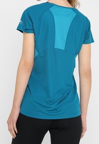 Kvinna som bär en kortärmad teal träningsskjorta med mesh-paneler på ryggen och svarta leggings, stående med ryggen vänd mot kameran.
