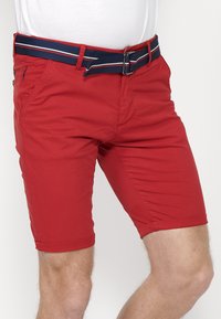 Shorts rouges à coupe ajustée, fabriqués en tissu léger. Ils comprennent des poches latérales, des revers roulés et une ceinture bleu marine avec des rayures blanches et rouges.