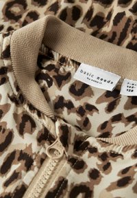 Leopardskinnsammet med nyanser av brunt och beige, med ett beige dragkedja, ribbad krage och en etikett som säger "basic needs by name it."