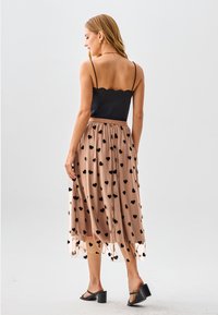 Schwarzes Top mit geschwungenem Saum, kombiniert mit einem nudefarbenen Rock mit schwarzen Herzmustern, transparentem Stoff und knöchellanger Schnitt, getragen zu schwarzen Sandalen mit Absatz.