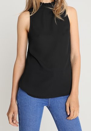 Femme portant un haut noir sans manches à col haut associé à un jean bleu en denim sur un fond clair uni.